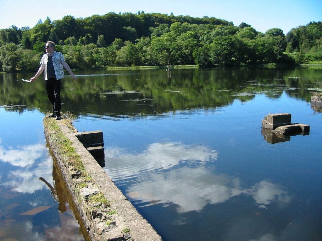 A man stands on a wall which juts out into a large pond. The pond is actually an outdoor swimming pool. In his hand, he holds a feather.